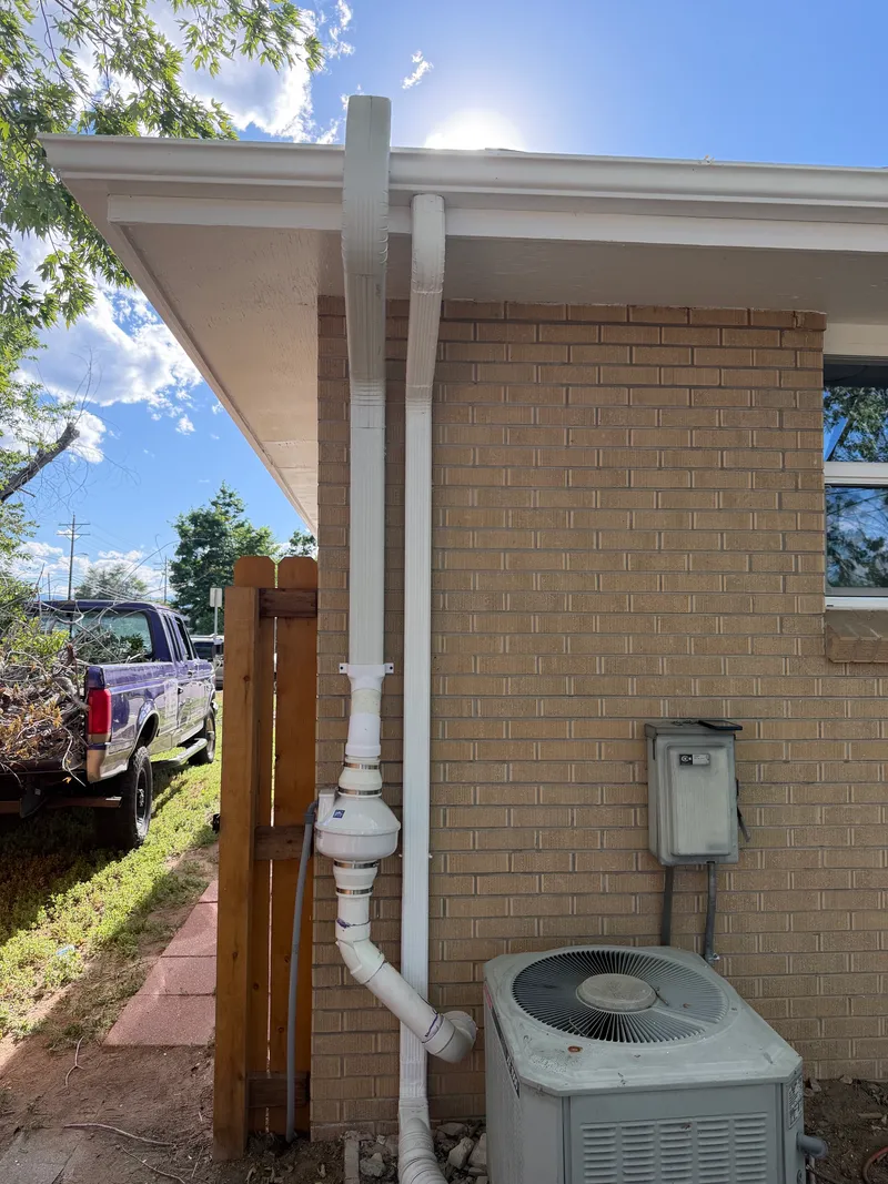 White radon fan and downspout run on buff brick, routed beside the AC unit